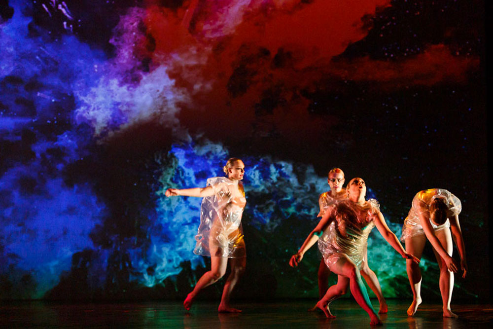 4 dancers wear clear plastic outfits as they dance in front of a galaxy projection in IM•MORTAL